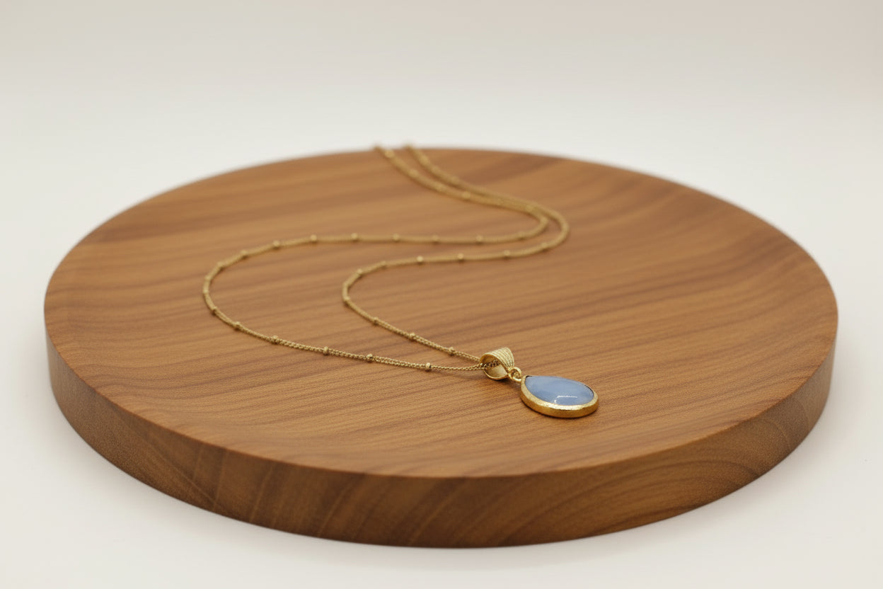 a necklace with gold chain and a blue stone pendant  is on a wooden round surface